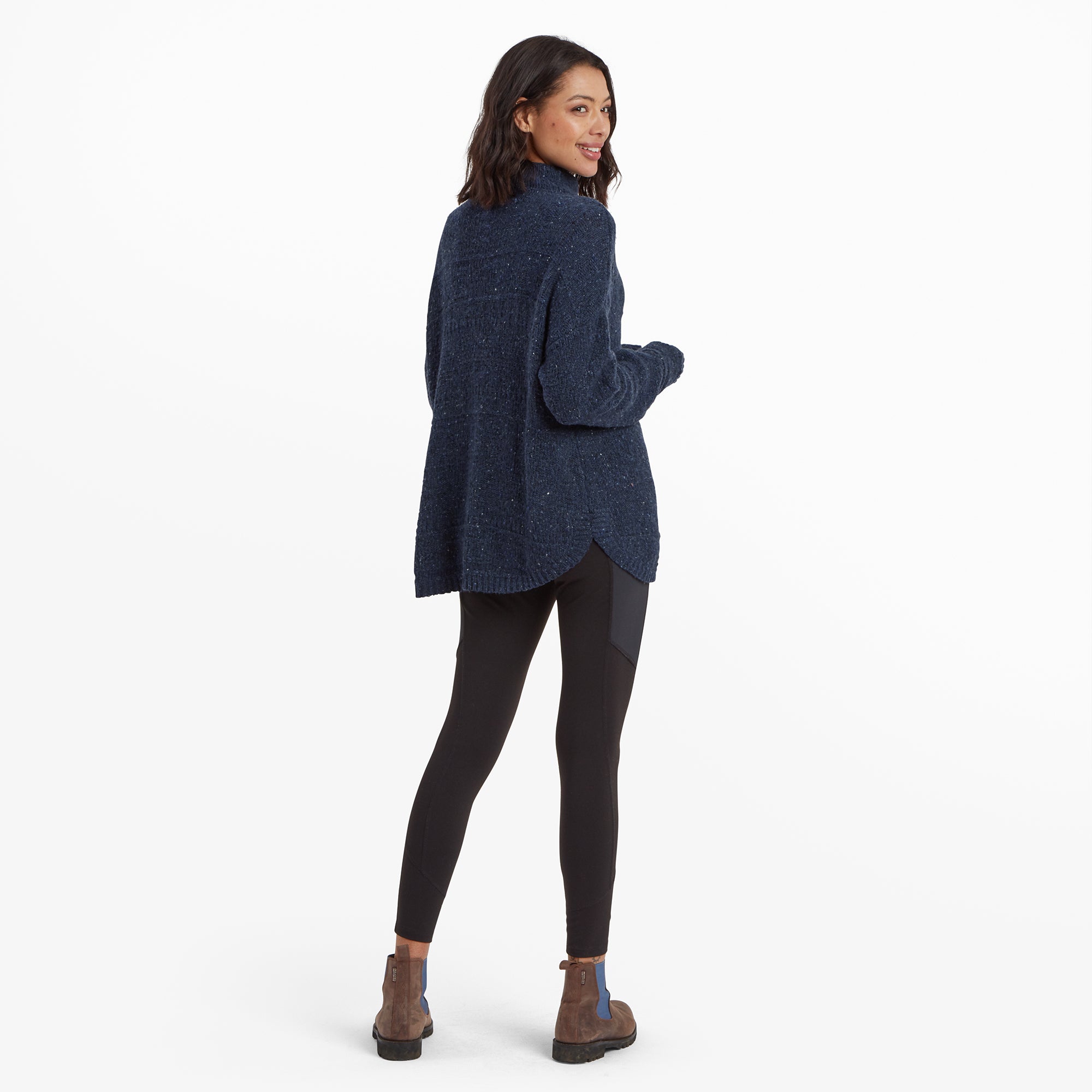 A back view of the model, showcasing the full length of the Sherpa Adventure Gear Yuden Pullover Sweater in Blue, which extends slightly past the hips. The model is turned slightly over the shoulder, smiling, and paired with leggings and stylish boots for a practical, outdoor-friendly look.