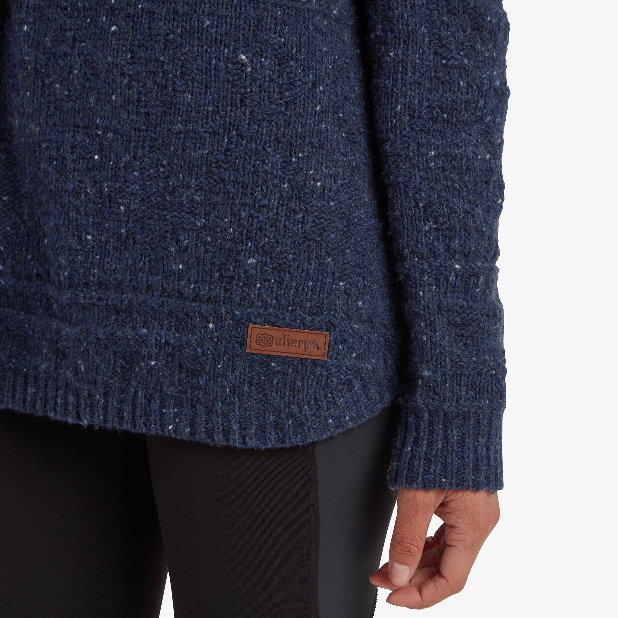 A close-up of the lower section of the Sherpa Adventure Gear Yuden Pullover Sweater in Blue, focusing on the Sherpa logo stitched onto a small leather patch. The curved hemline and detailed ribbed knit are also highlighted, paired with the edge of the black leggings for a cohesive look.