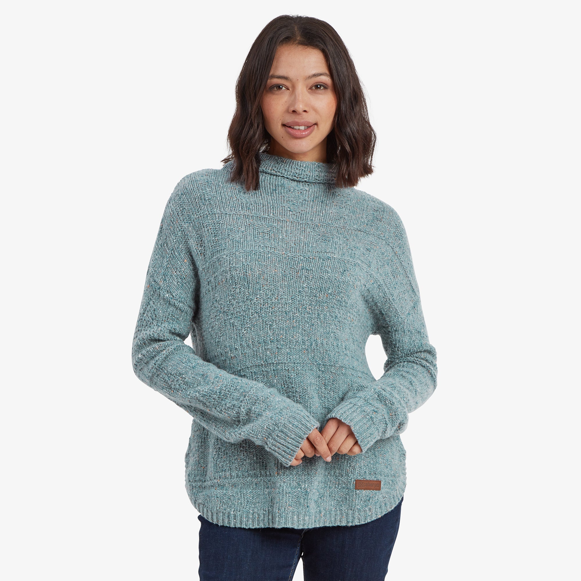 A woman wearing a soft, textured Sherpa Adventure Gear Yuden Pullover Sweater in Green with a relaxed high neckline. The sweater has a loose fit, slightly longer hem at the back, and full-length sleeves. She pairs it with dark blue jeans, standing casually with a gentle smile.