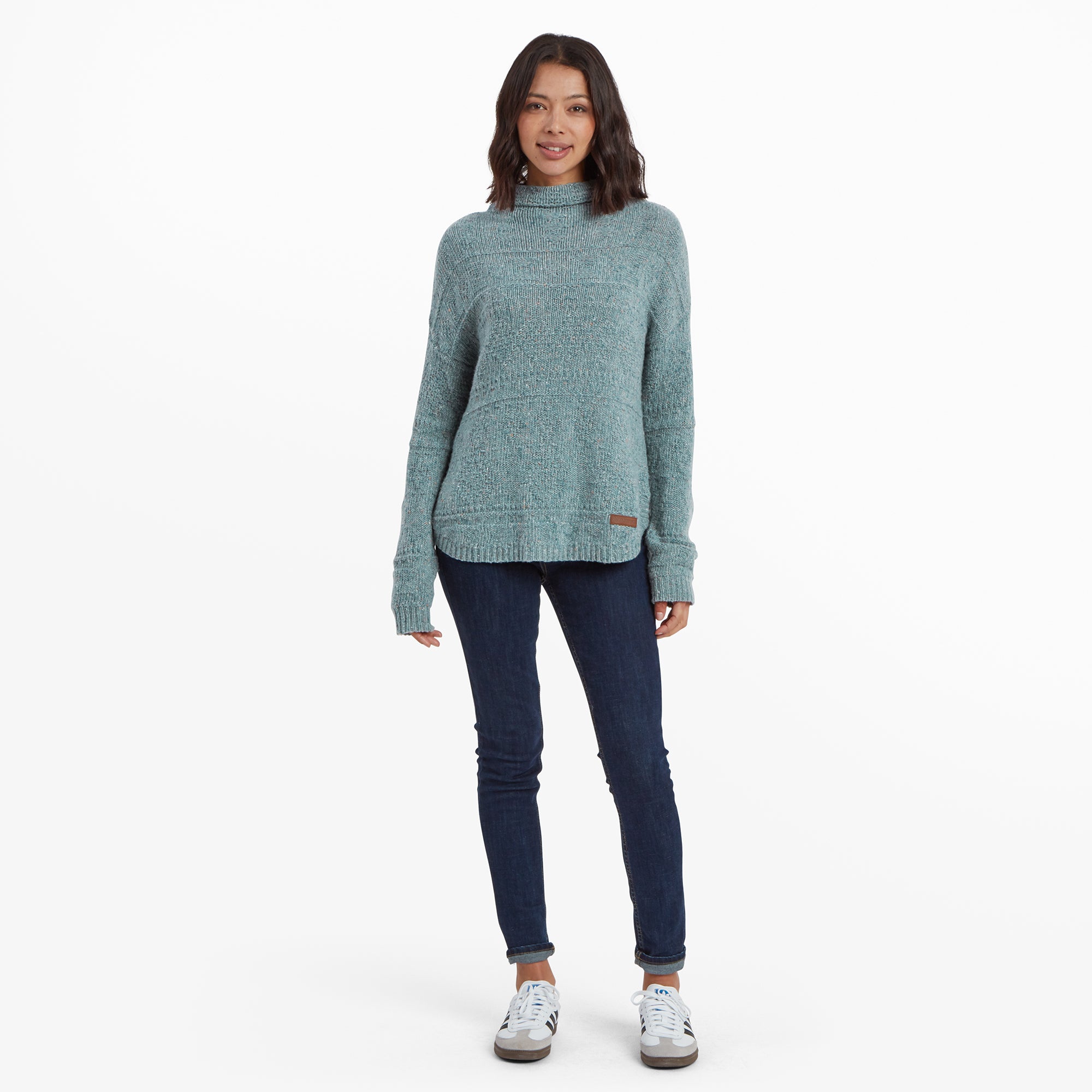 The same woman showcasing the full outfit, emphasising the relaxed and slightly oversized fit of the Sherpa Adventure Gear Yuden Pullover Sweater in Green. She pairs it with slim-fit jeans and white trainers, standing straight with hands by her sides.