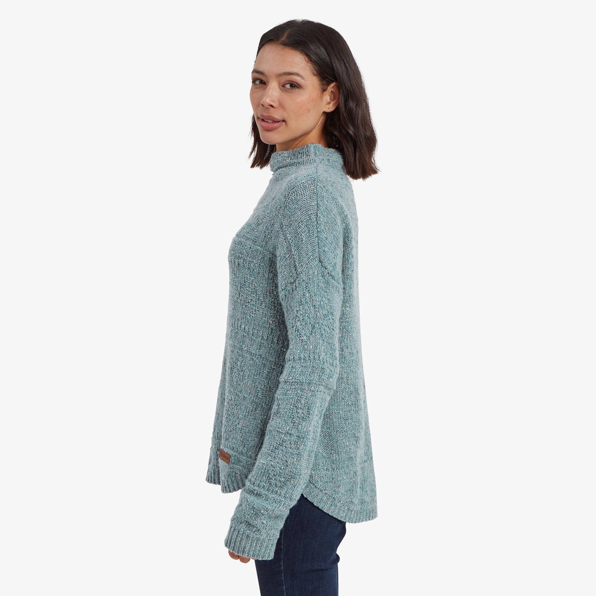 A side view of the model in the Sherpa Adventure Gear Yuden Pullover Sweater in Green, showcasing the slight curve of the hemline that is longer at the back. The textured knit and loose fit of the sweater are visible, paired with her jeans for a stylish yet cozy look.