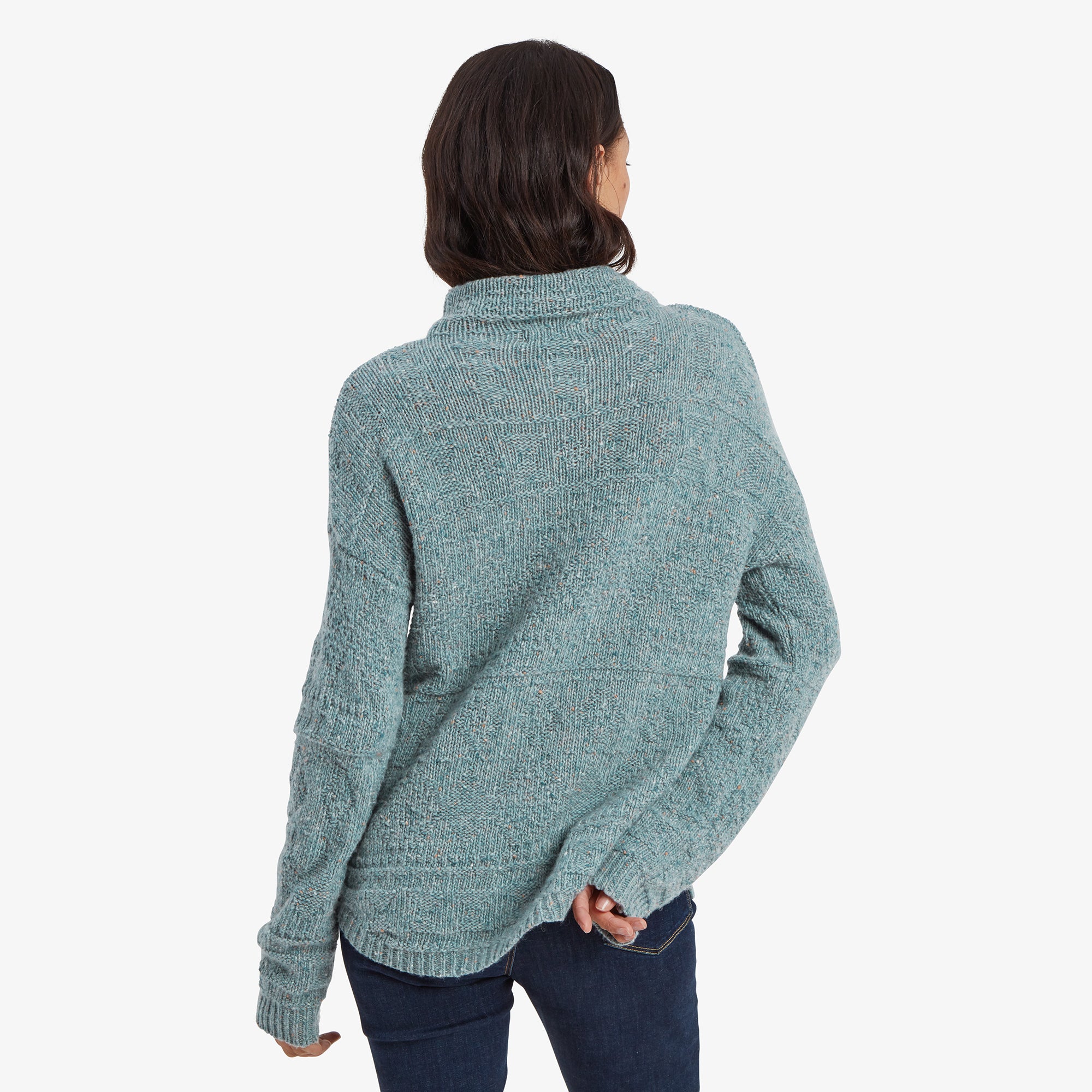 A rear view of the Sherpa Adventure Gear Yuden Pullover Sweater in Green, highlighting its slightly longer hemline and seamless back design. The high neckline is folded softly at the back, while the loose fit complements her jeans for a casual appearance.