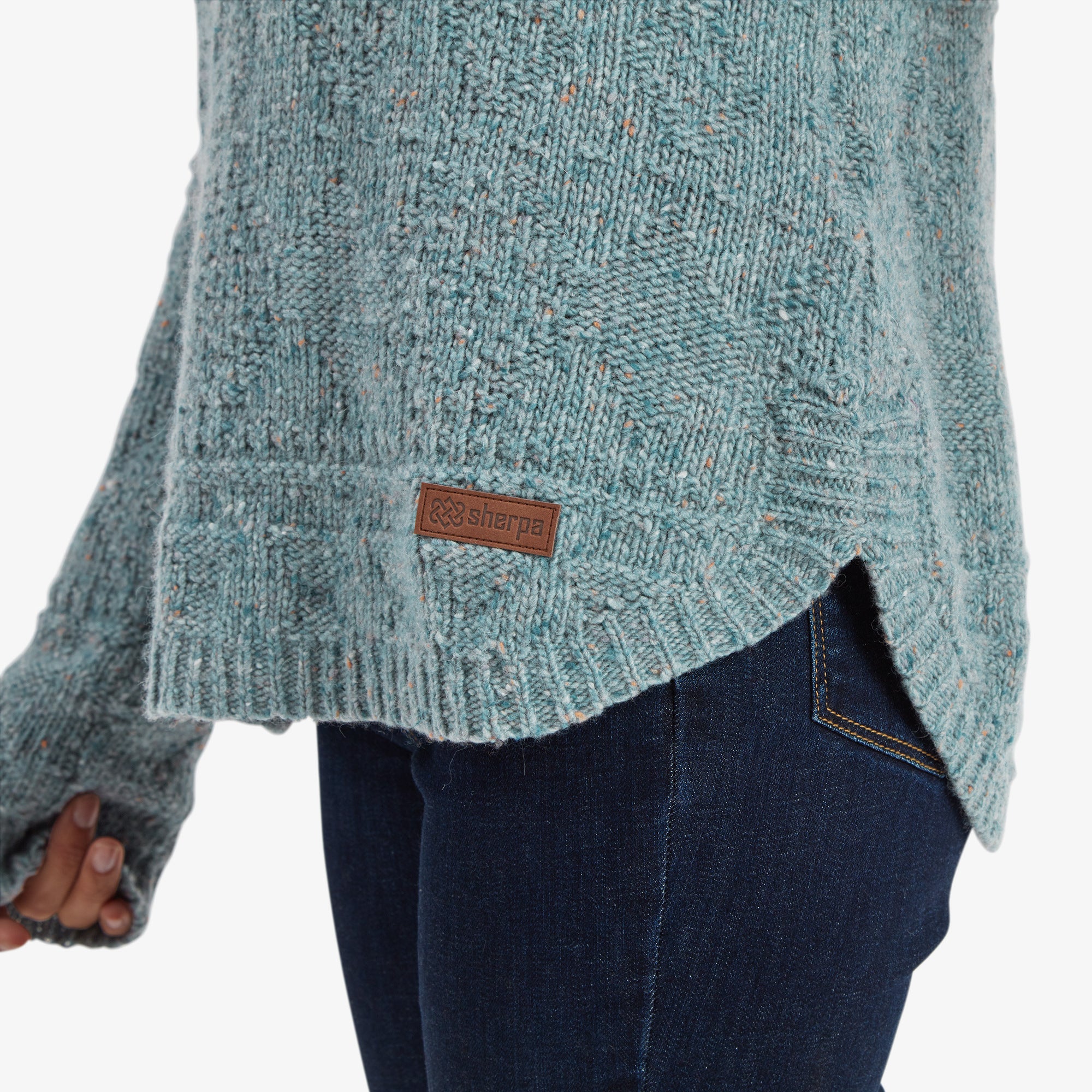 A detailed view of the Sherpa Adventure Gear Yuden Pullover Sweater in Green’s hem, showing its textured knit design and the small brown Sherpa logo patch on the side. The curved hemline pairs perfectly with the dark blue jeans for a modern and casual look.