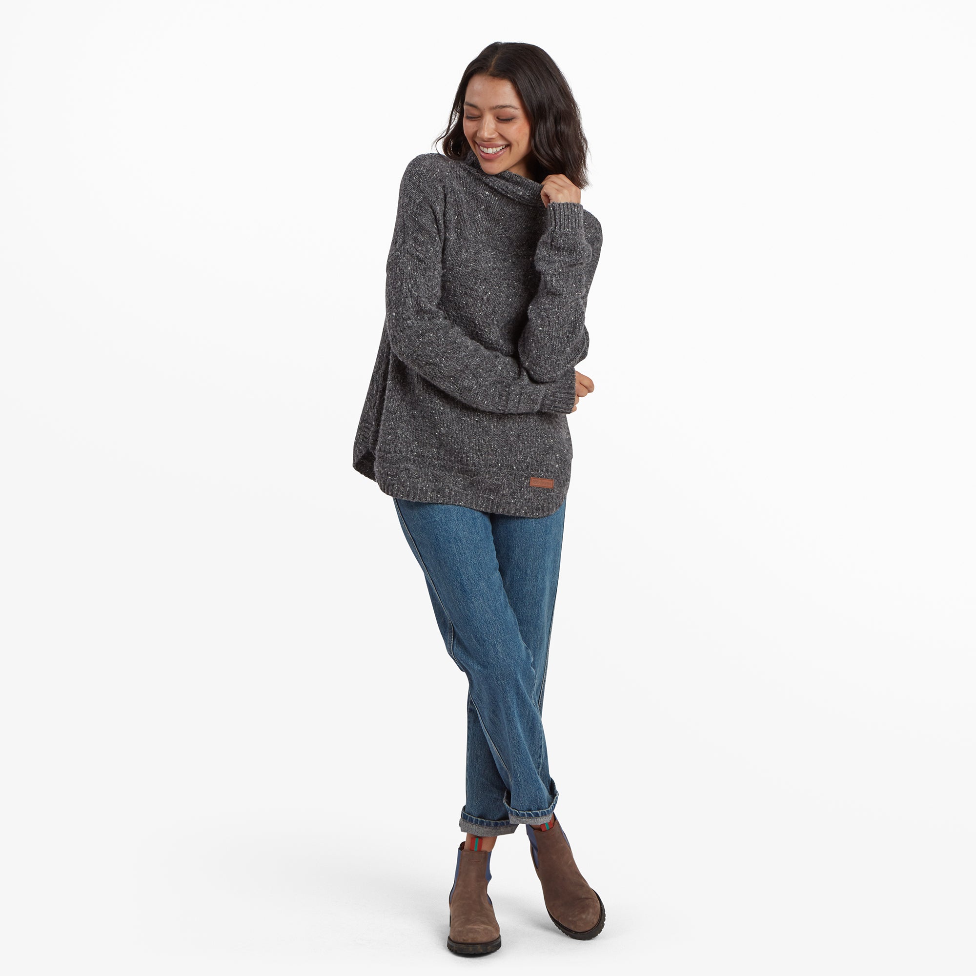 A full-body shot of the woman dressed in the Sherpa Adventure Gear Yuden Pullover Sweater in Grey. She stands in a casual pose, holding the collar of the sweater with a smile. The outfit is styled with blue jeans and brown ankle boots, giving a cozy and casual vibe.