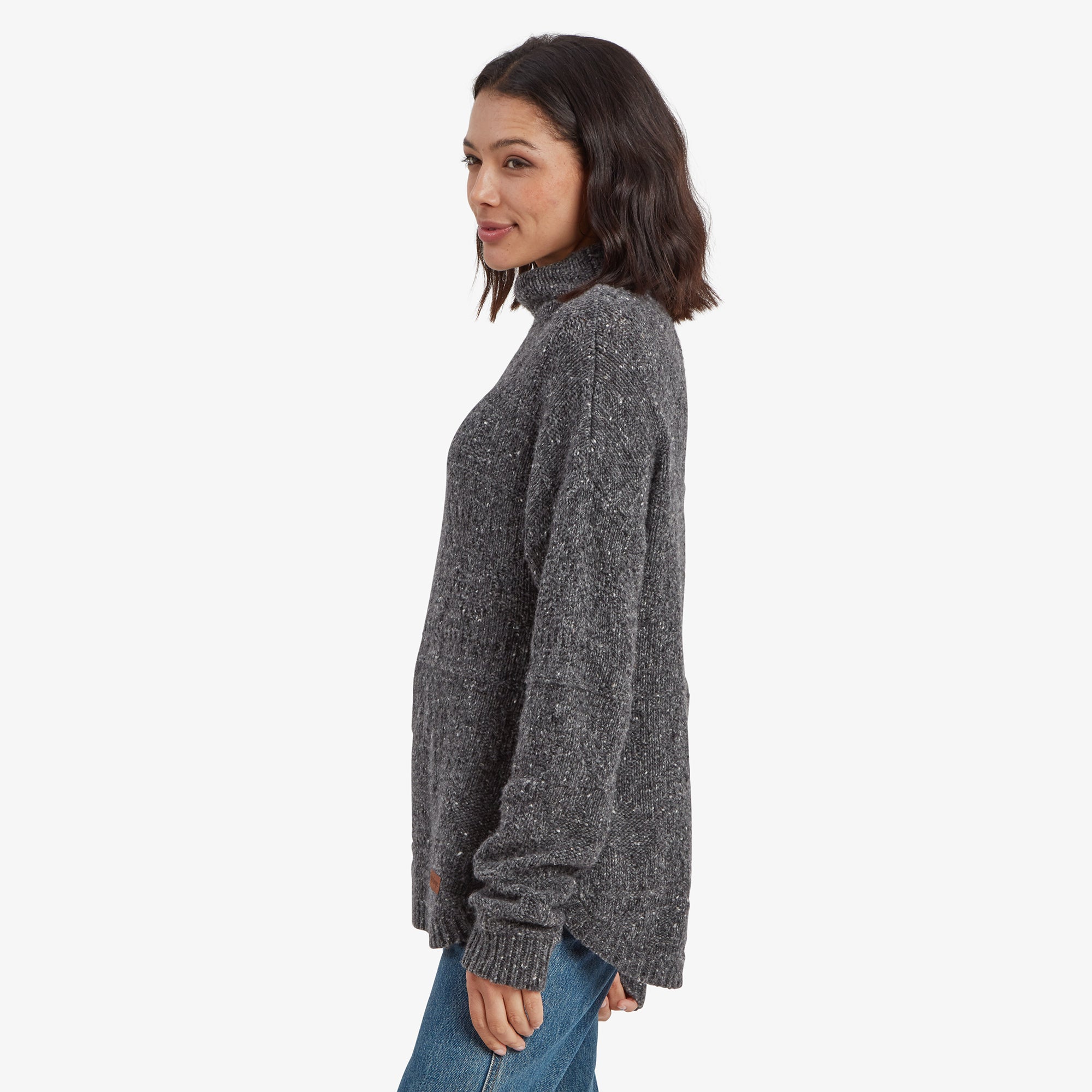 A side profile of the Sherpa Adventure Gear Yuden Pullover Sweater in Grey, highlighting its longer hemline and slightly loose silhouette. The textured knit pattern is visible, and the high neckline adds a warm and snug look. The woman looks over her shoulder, giving a soft smile.