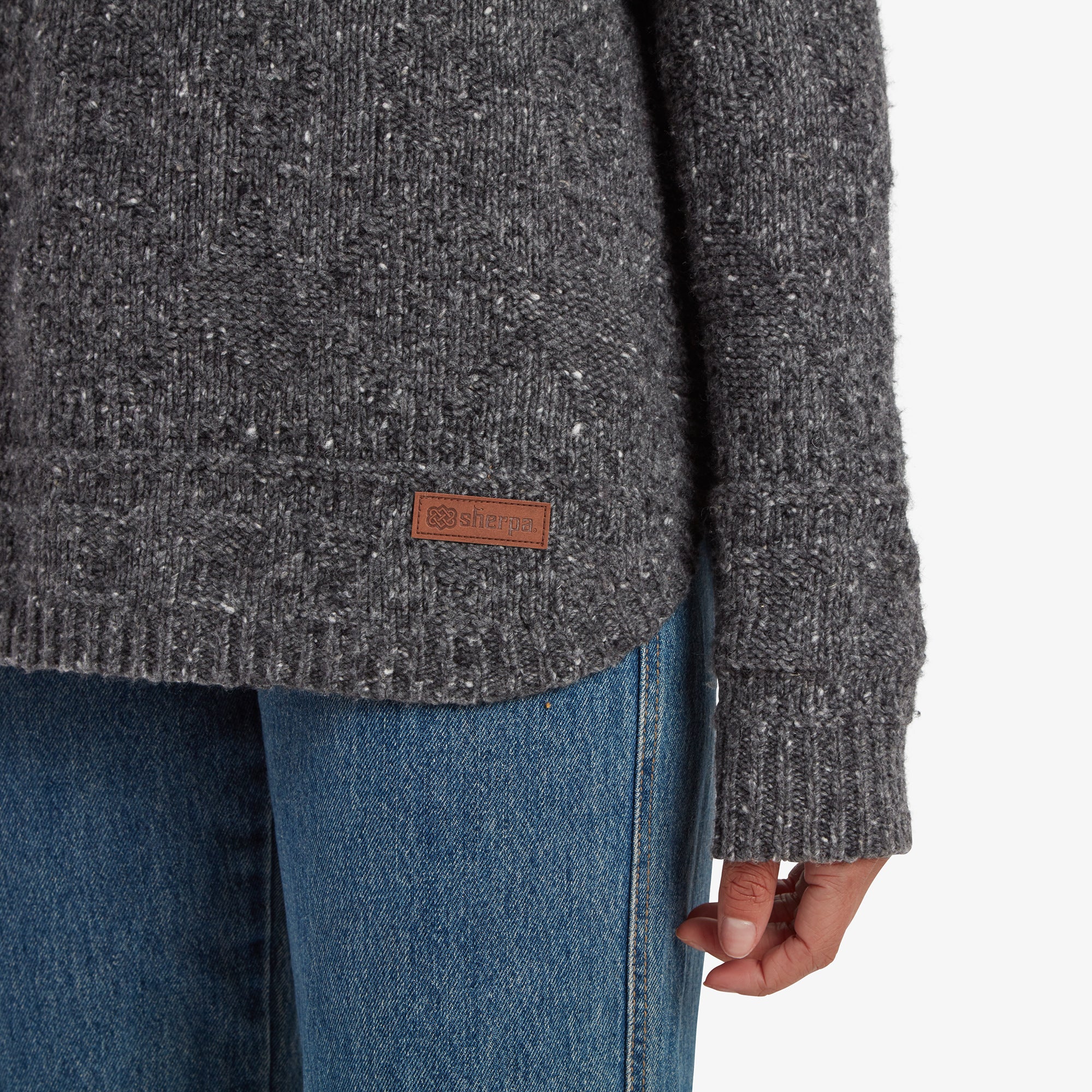 A close-up detail of the lower hemline on the Sherpa Adventure Gear Yuden Pullover Sweater in Grey, showing the ribbed edge and a small leather tag with the Sherpa logo. The textured knit pattern continues throughout, paired with the blue jeans for a casual look.