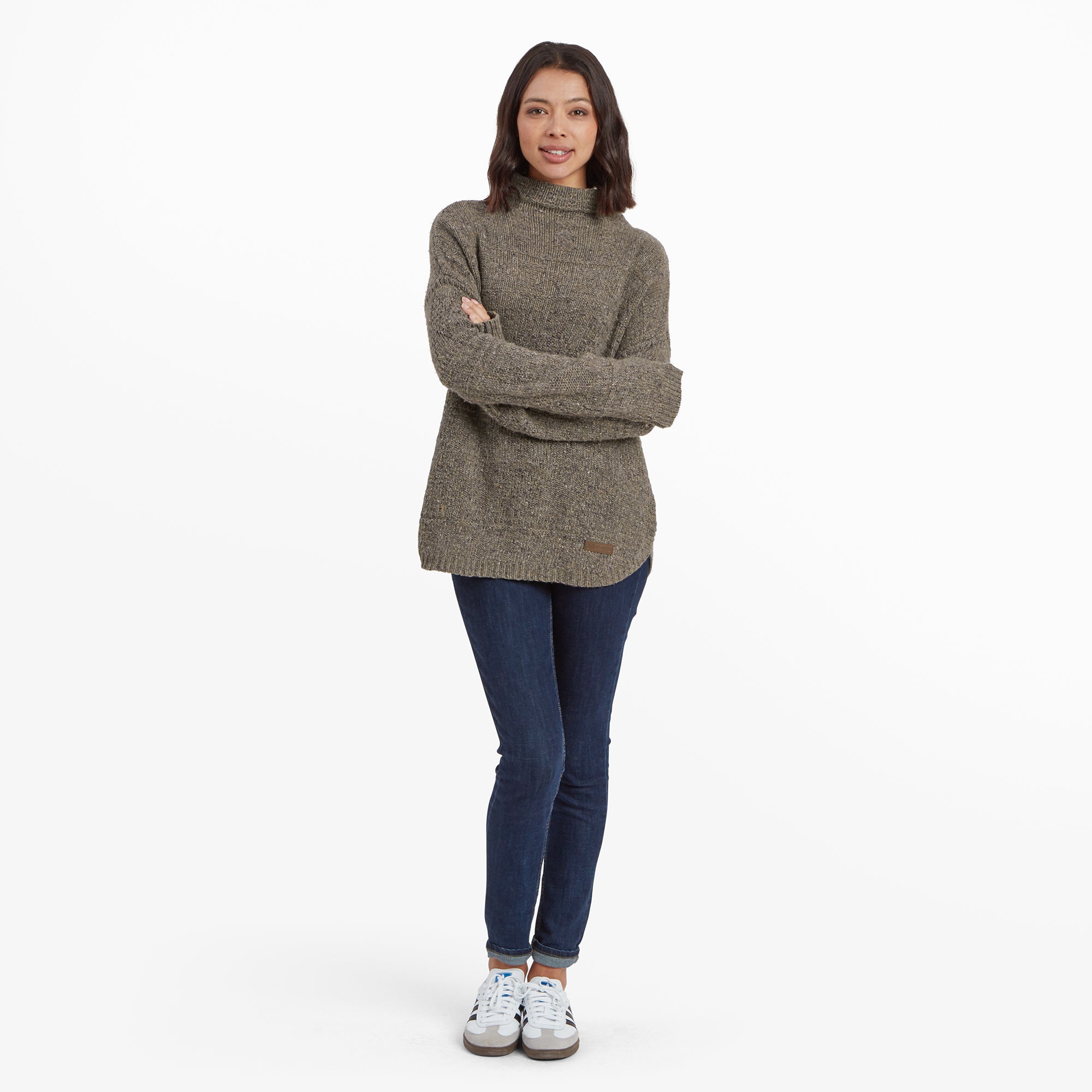 A full-length view of a woman wearing a Sherpa Adventure Gear Yuden Pullover Sweater in Grey with a high neckline, dark blue jeans, and white trainers. She is standing with arms crossed, smiling softly. The sweater’s relaxed fit and rounded hem are clearly visible.