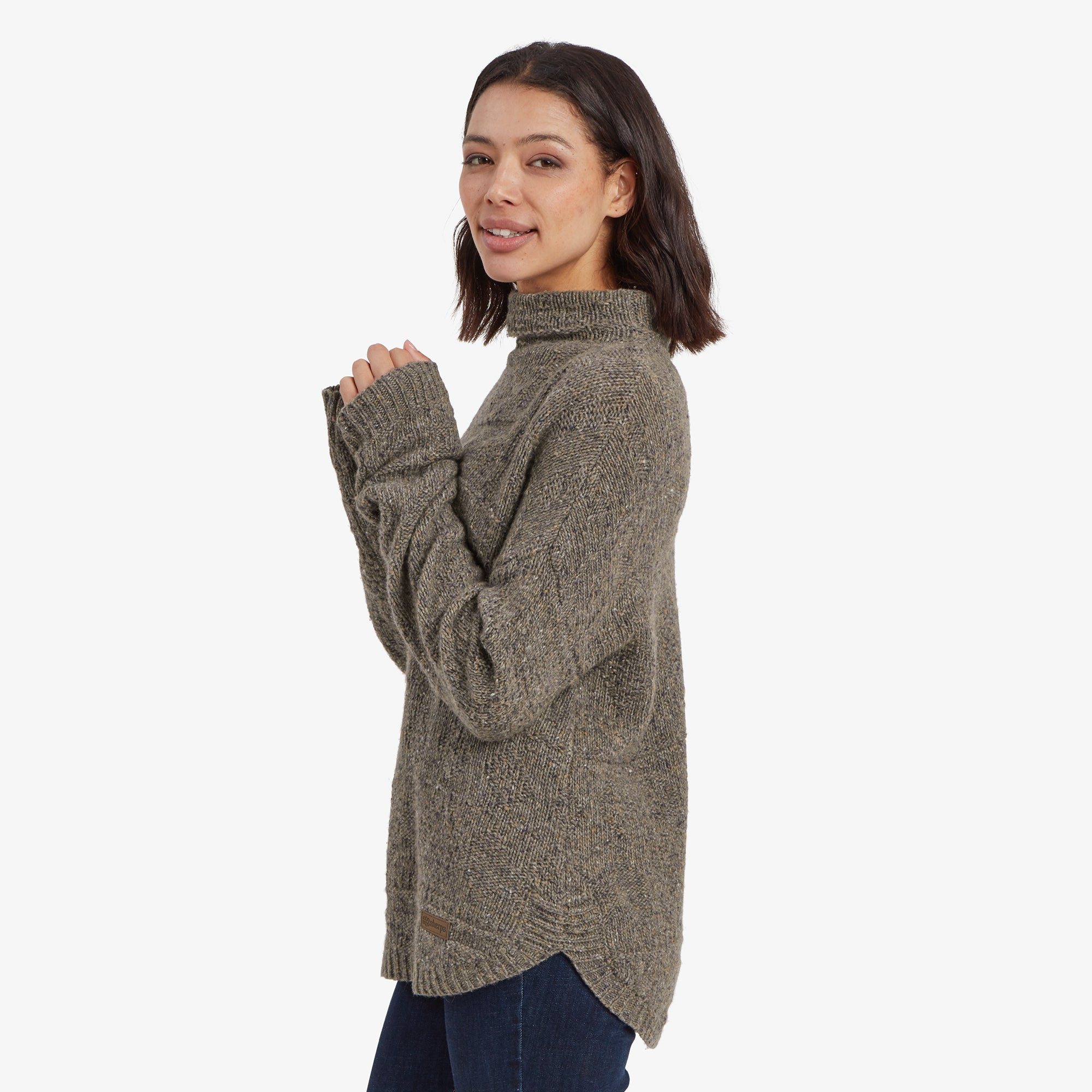 A side profile of a woman wearing a Sherpa Adventure Gear Yuden Pullover Sweater in Grey. The high neckline and textured knit detail are highlighted as she gently folds her hands close to her chest. The sweater's loose fit and curved hem provide a cozy and stylish appearance.