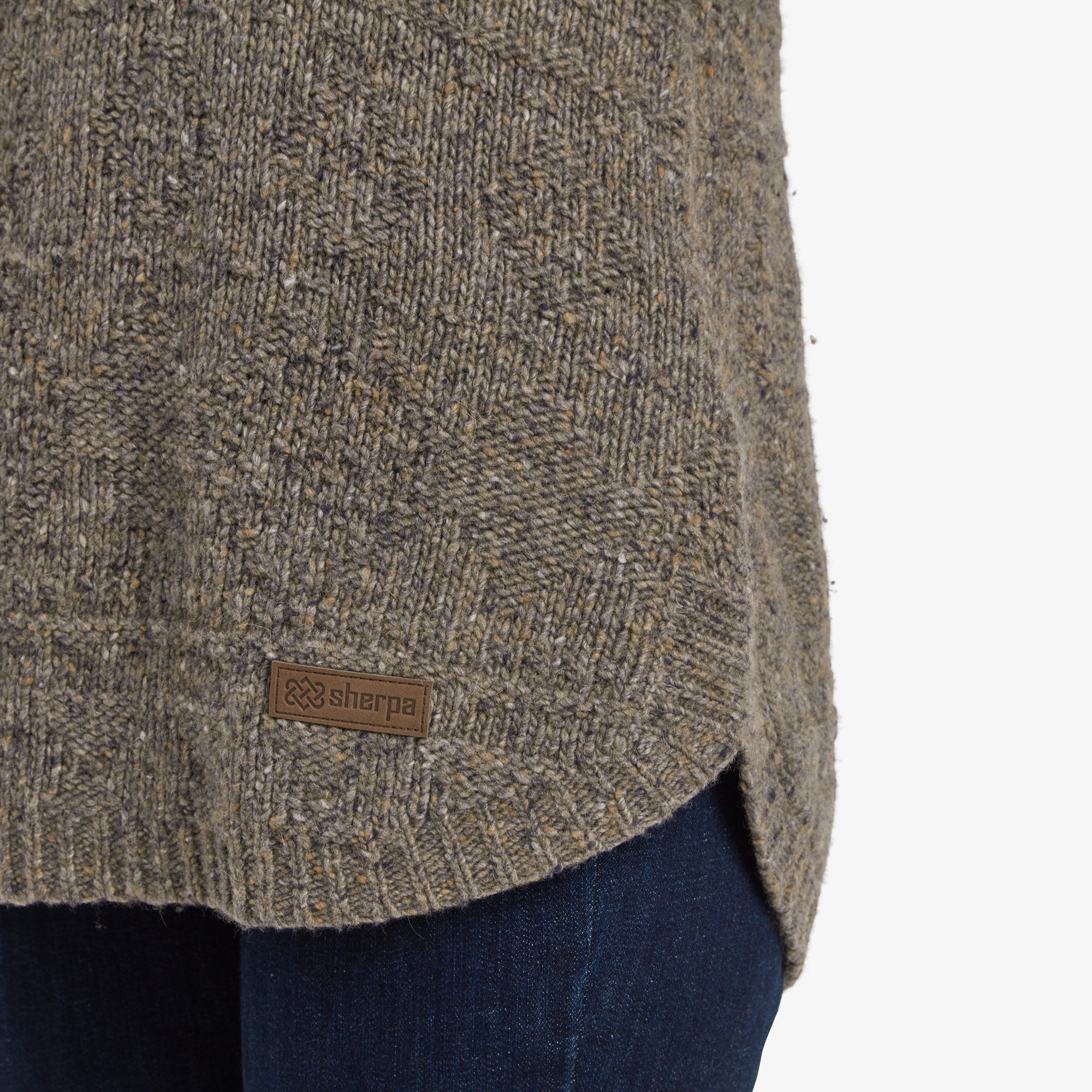 A close-up of the bottom hem of the Sherpa Adventure Gear Yuden Pullover Sweater in Grey, showing a small brown Sherpa logo tag sewn on the side. The curved hem and the texture of the fabric are clearly visible, paired with dark blue jeans.