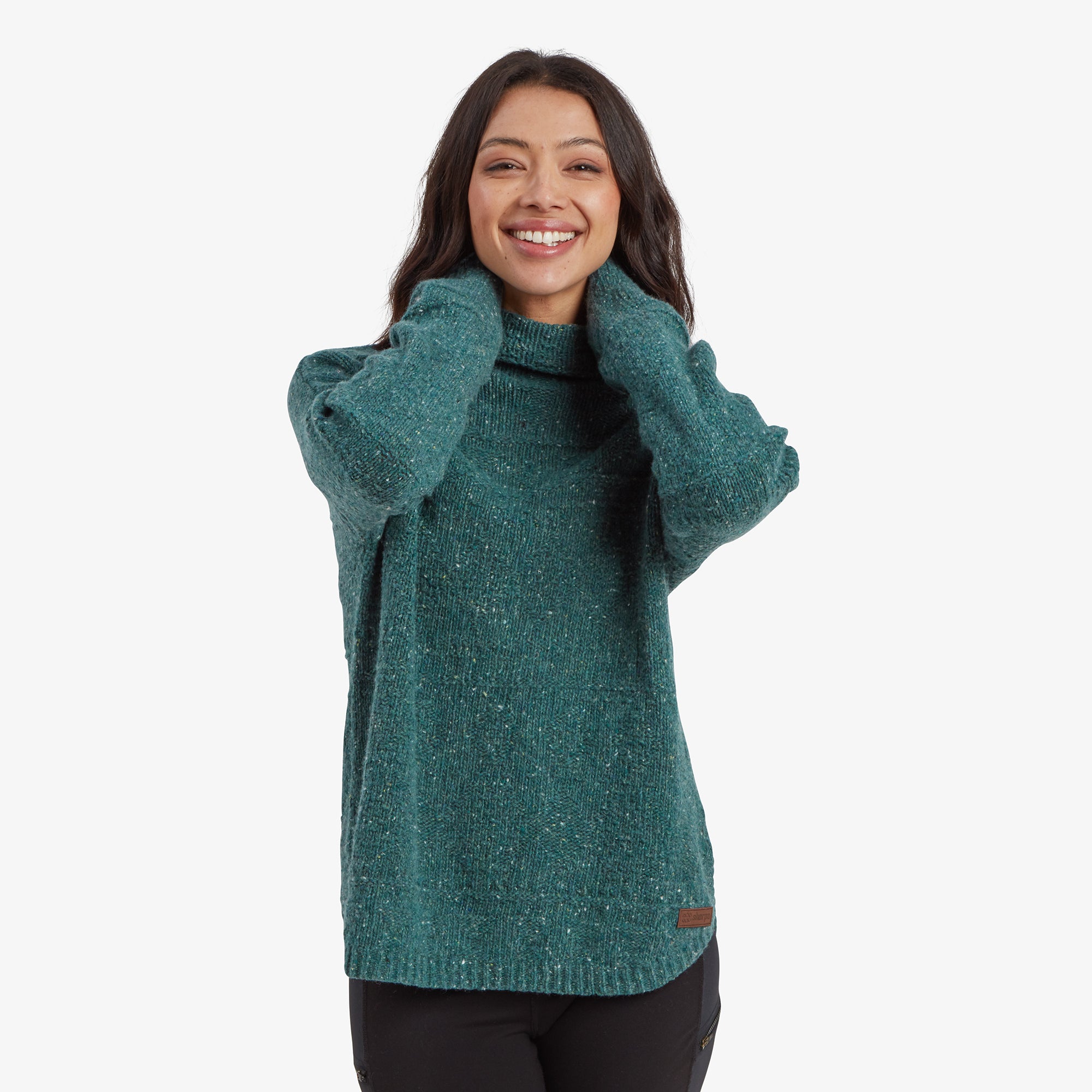 A woman with long, dark brown hair and a bright smile wears a Sherpa Adventure Gear Yuden Sweater in Blue with a relaxed fit. She gently holds the collar of the sweater with both hands, pulling it up slightly. The sweater has a speckled knit pattern with subtle variations in color. She is also wearing black leggings, and the background is a clean white.