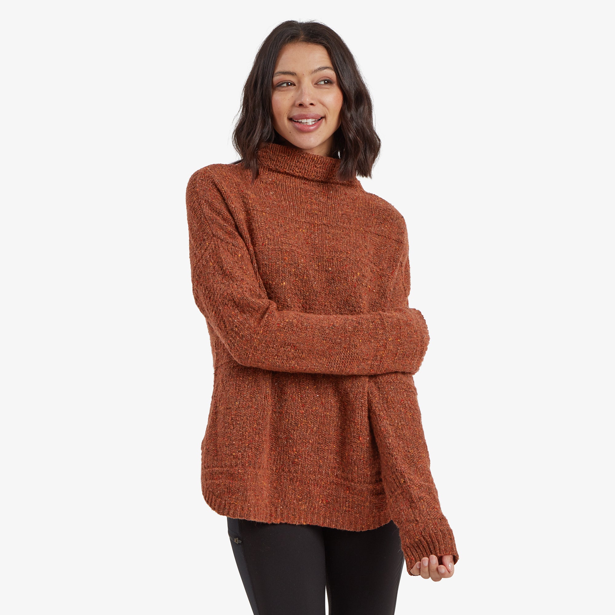 A close-up front view of a woman wearing a loose-fitting Sherpa Adventure Gear Yuden Sweater in Brown. The texture of the sweater reveals a mix of earthy tones with subtle speckles of orange and yellow, complemented by a relaxed fit. The model smiles softly, with her hands playfully adjusting the collar.