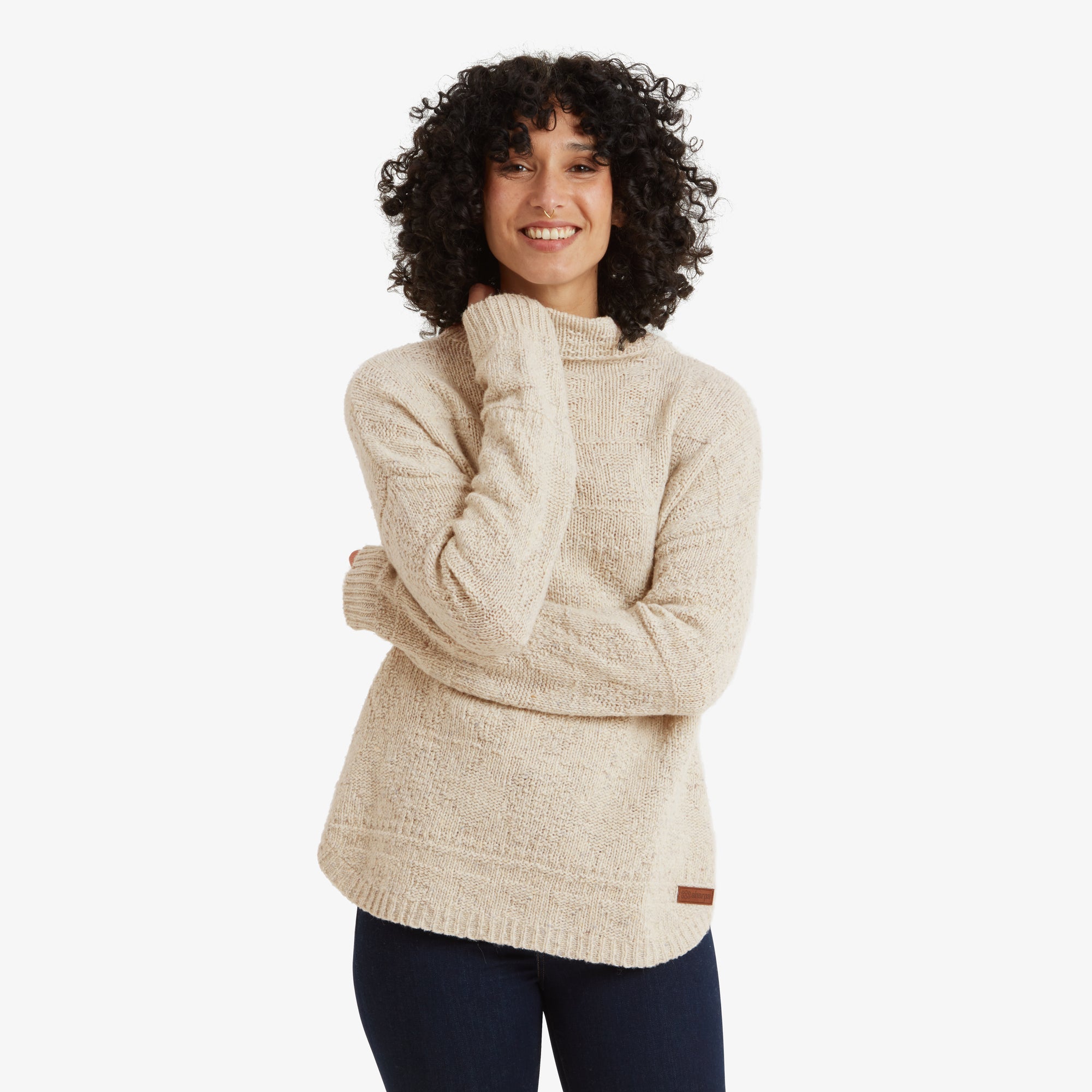 A front-facing image of a woman wearing a cozy Sherpa Adventure Gear Yuden Sweater in White with a high neckline, textured knit pattern, and relaxed fit. She is smiling warmly with her hand gently resting near her chin, highlighting the comfortable and stylish design of the sweater.