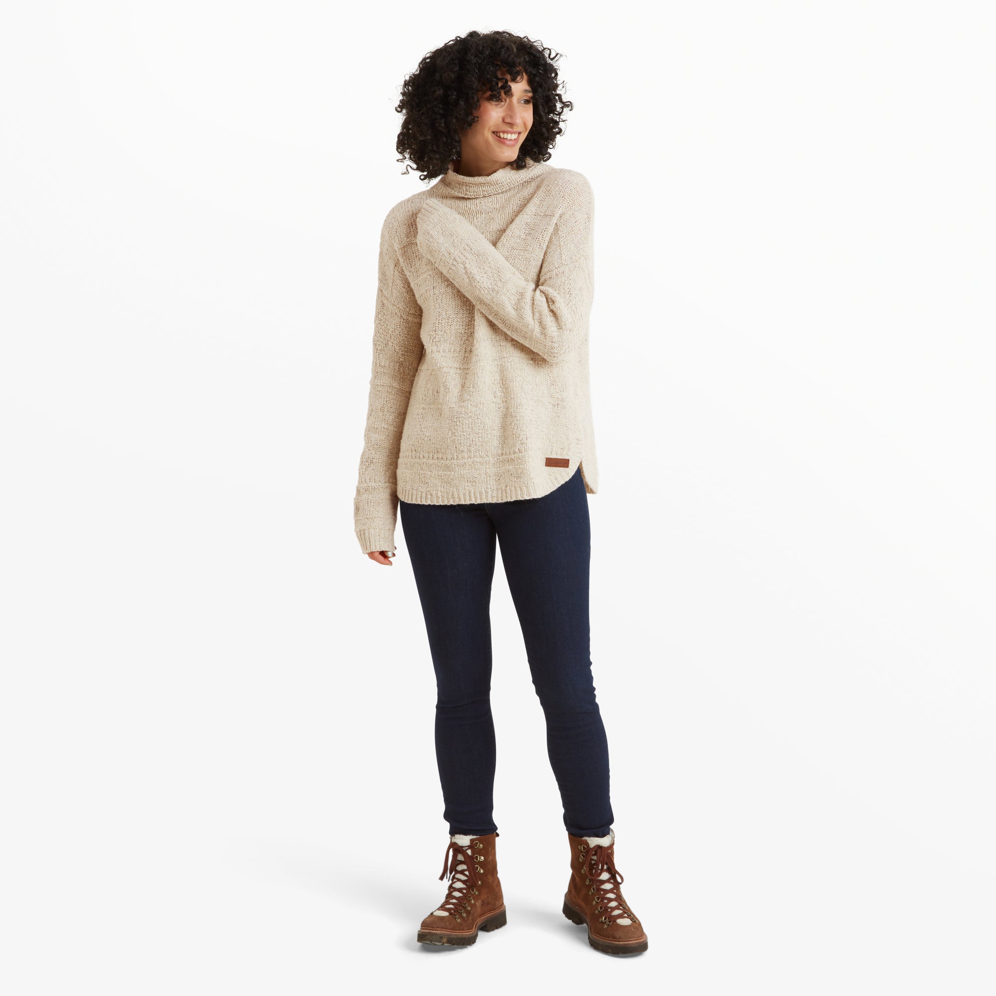 The woman is standing in a relaxed posture, showcasing the Sherpa Adventure Gear Yuden Sweater in White paired with dark blue skinny jeans and brown lace-up boots. The overall outfit demonstrates the casual and warm styling perfect for colder weather.