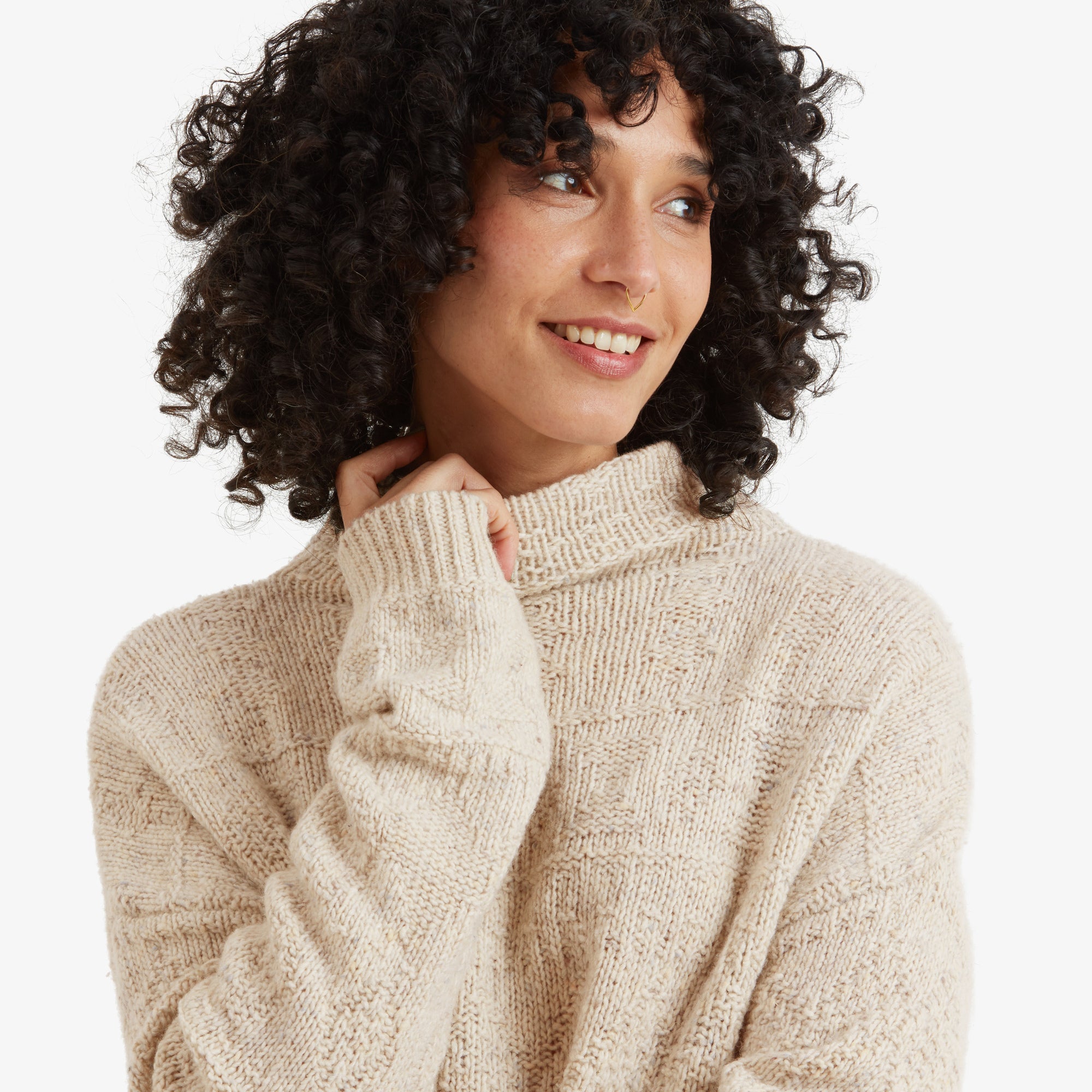 A zoomed-in shot of the woman smiling with her hand near her face, showing the intricate knit texture of the high neckline and upper chest area on the Sherpa Adventure Gear Yuden Sweater in White.