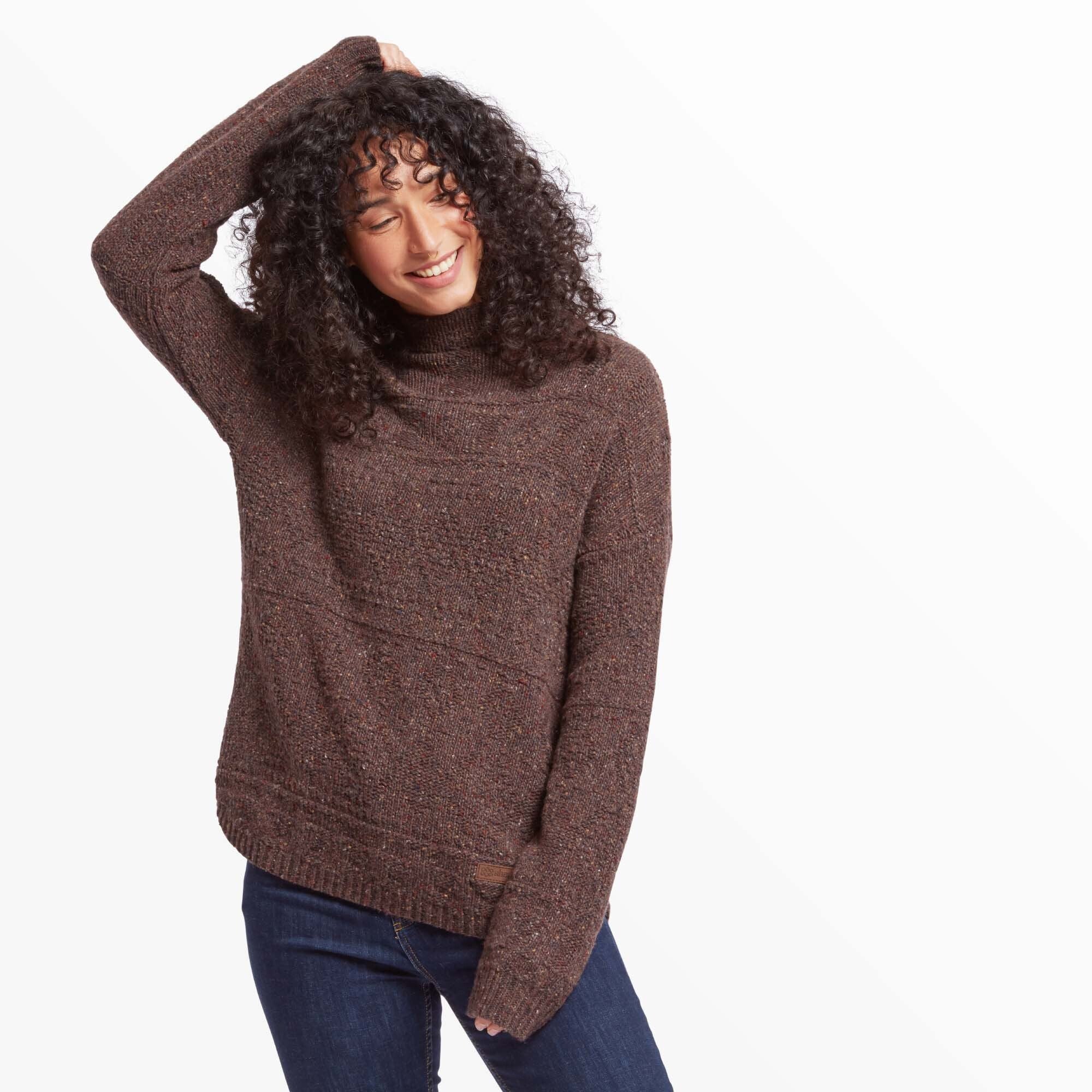 A woman is modeling a relaxed Sherpa Adventure Gear Yuden Sweater in Red with intricate knit patterns. She poses with one hand resting on her head, smiling with her eyes closed. The sweater features subtle speckles of colour throughout and a slight asymmetrical hem. She pairs it with fitted dark blue jeans.