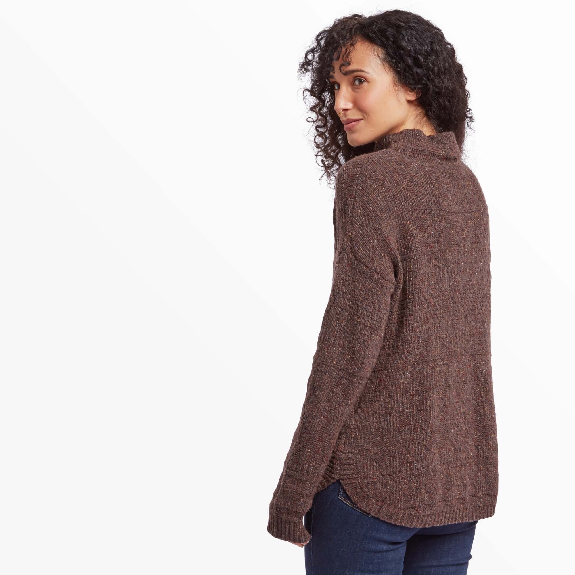 A side and back view of the Sherpa Adventure Gear Yuden Sweater in Red as worn by the same model. The hem features a slight curve with a ribbed texture, and the knit detailing on the back is visible. She turns her head slightly over her shoulder with a soft expression, her curly hair cascading down.