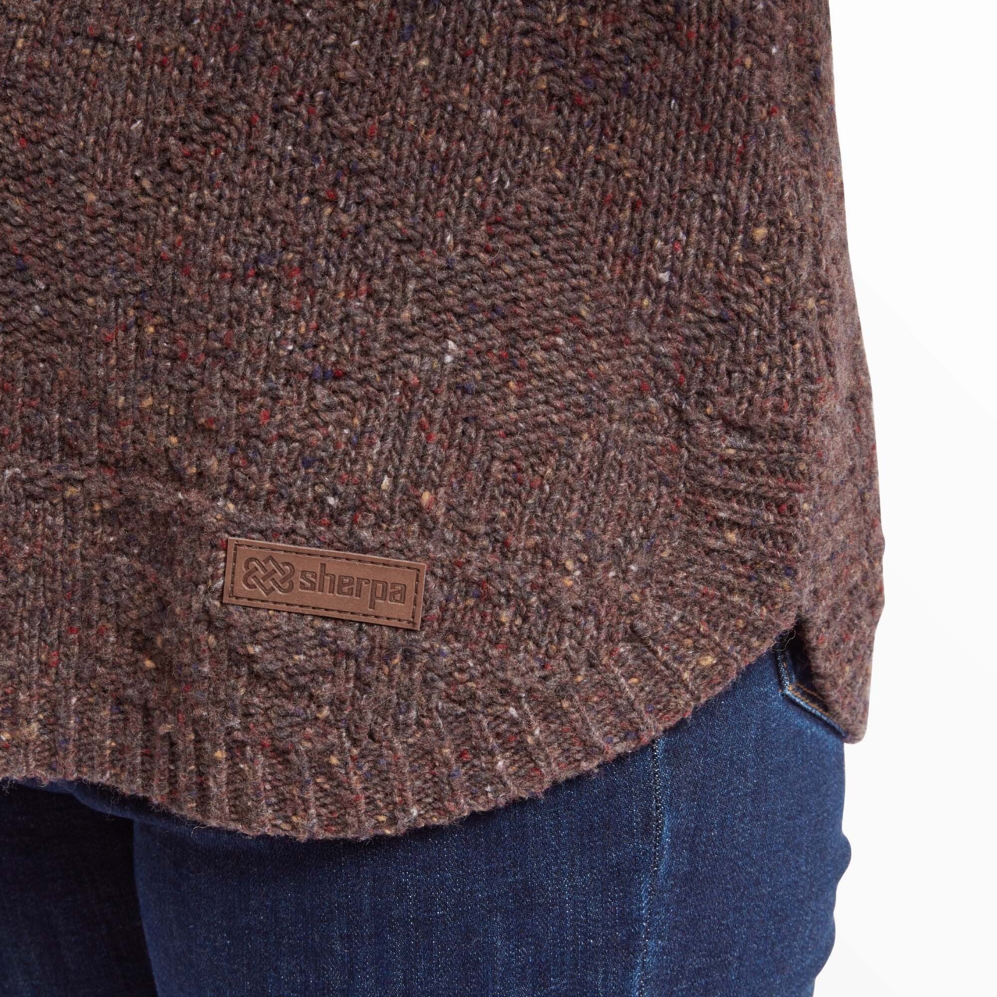 A close-up of the lower hem of the Sherpa Adventure Gear Yuden Sweater in Red, showcasing the textured knit pattern and a small brown leather Sherpa logo patch. The hemline curves gently over a pair of dark blue jeans. The speckled detailing of the fabric is emphasised in this image.